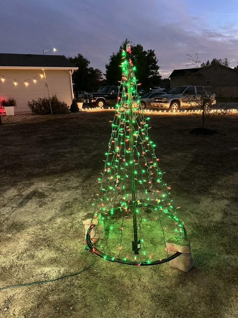 Custom cone tree wrapped in green and red lights in a yard at dusk